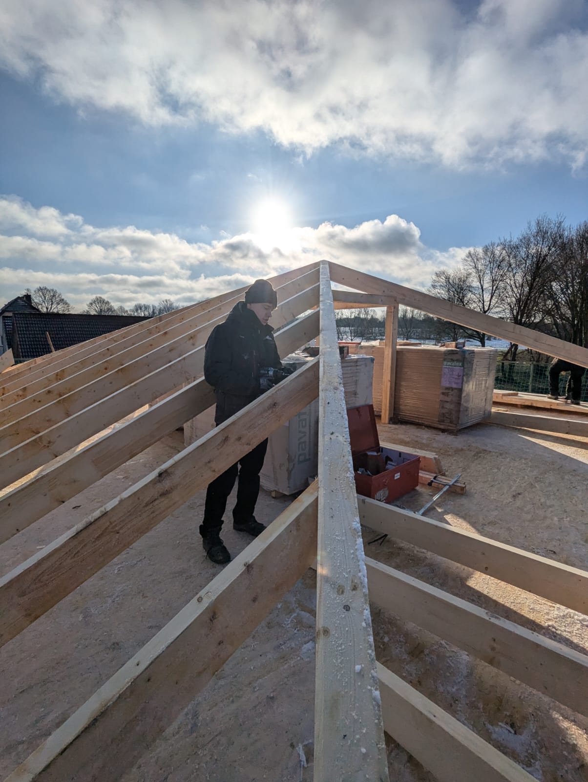Zimmerer der Zimmerei Tragwerk Pietsch arbeitet auf einem frisch gestellten Dachstuhl im Gegenlicht – Sparren und First sichtbar, Neubau-Baustelle in Steinhagen, Kreis Gütersloh
